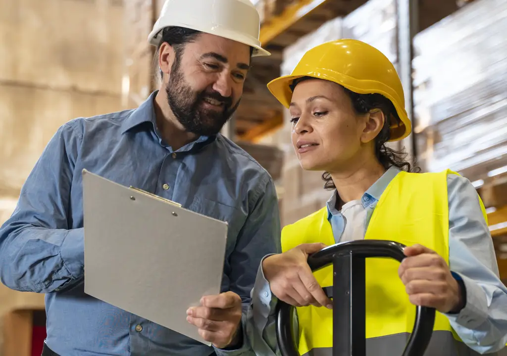 Two warehouse workers looking at a clipboard. Both are wearing a hard hat and one is wearing a high-vis vest