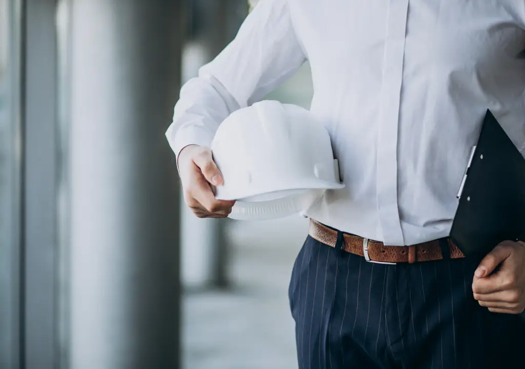 The mid-section of a man wearing smart clothes, holding a hard hat and a clip board